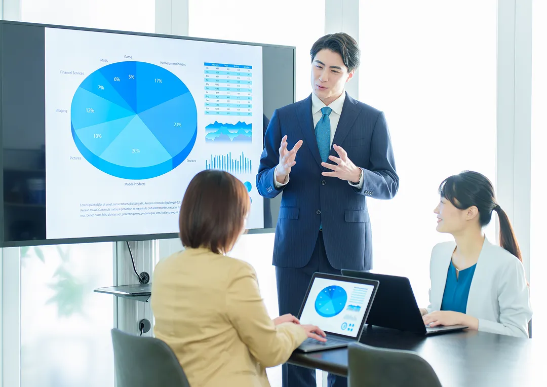 スーツ姿の男性が、大型ディスプレイに表示された円グラフを使ってプレゼンをしている。