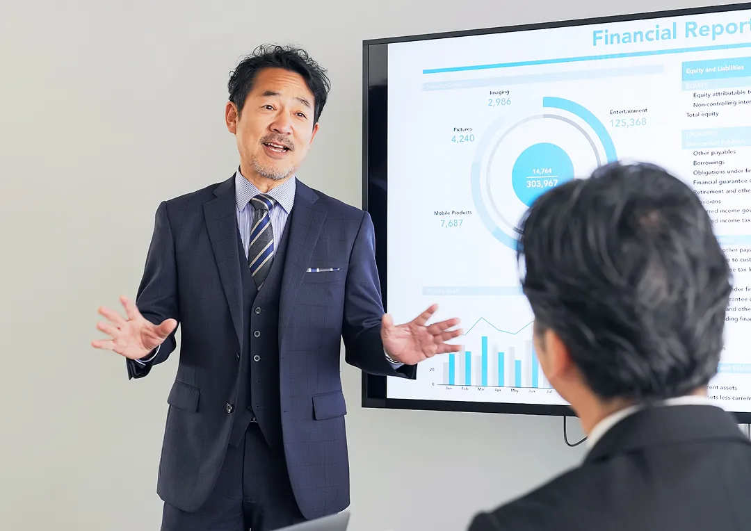 スーツ姿の中年男性が、財務報告のスライドを使ってプレゼンテーションをしている。