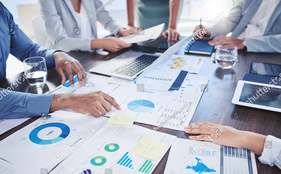 会議テーブルに紙の資料とノートPCが並び、参加者が資料を見ながら話し合っている様子。